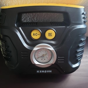 May include: A black and yellow Kensun portable air compressor featuring a top handle, two yellow buttons labelled AC and DC, and a central analogue pressure gauge with a silver rim. The device has a moulded plastic housing with side vents.