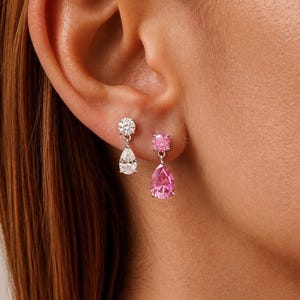 May include: Two drop earrings featuring a metallic setting. One earring has a clear round stud with a clear pear-shaped drop. The other earring has a pink round stud with a pink pear-shaped drop. Both earrings are secured with a post back.