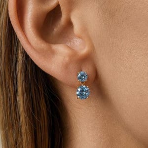 May include: A gold drop earring featuring two round, light blue faceted gemstones set in a vertical arrangement. The smaller stone is positioned at the top, while the larger stone hangs below, both secured by gold prongs against the earlobe.