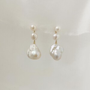 May include: A pair of earrings with a gold-coloured setting, each featuring a combination of small, round pearls and large, irregularly shaped baroque pearls. The pearls are creamy white in colour and hang from a delicate chain.