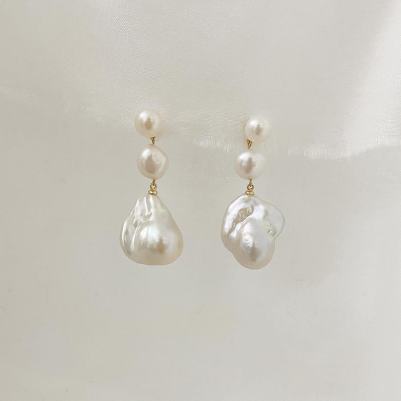 May include: A pair of earrings with a gold-coloured setting, each featuring a combination of small, round pearls and large, irregularly shaped baroque pearls. The pearls are creamy white in colour and hang from a delicate chain.