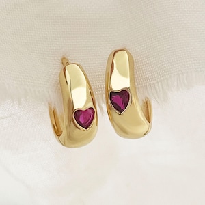 May include: A pair of gold hoop earrings with a small red heart-shaped gemstone in the center of each hoop.
