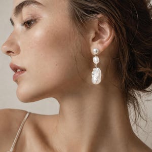 Puede incluir: Un pendiente colgante con tres perlas barrocas blancas de diferentes tamaños dispuestas verticalmente. La perla superior es pequeña y redonda, la del medio es de tamaño mediano e irregular, y la inferior es la más grande con superficie texturizada.