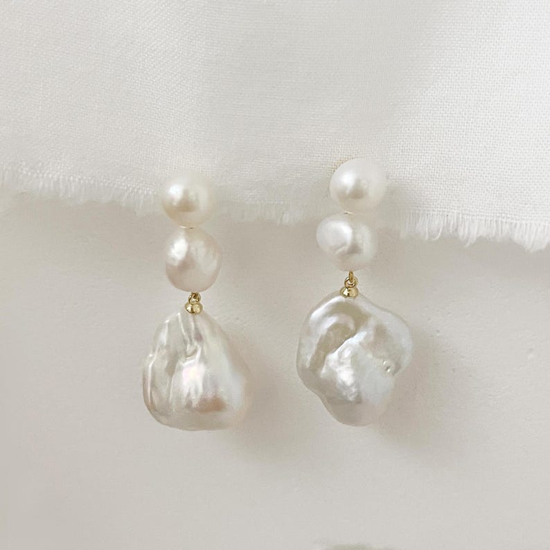 May include: A pair of pearl drop earrings. Each earring has three white pearls, with a large, irregularly shaped pearl at the bottom. The pearls are connected by gold-coloured metal. The earrings are set against a white background.