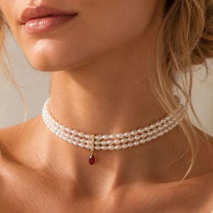 Puede incluir: Un collar tipo gargantilla de tres hilos con pequeñas perlas blancas ovaladas. Una barra vertical dorada engastada con diminutas piedras transparentes une los hilos en el centro, suspendiendo un colgante de piedra roja en forma de lágrima.