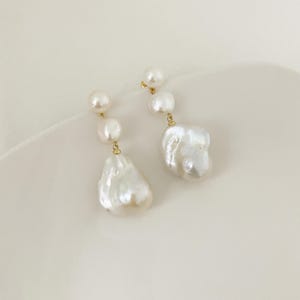 May include: A pair of earrings with three white pearls of different sizes hanging from a gold-coloured post. The largest pearl has an irregular, baroque shape. The earrings are displayed on a white surface.