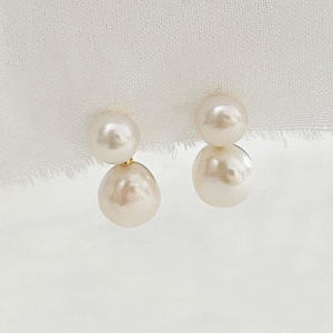May include: A pair of pearl earrings. Each earring has a small, round pearl above a larger, teardrop-shaped pearl. The pearls are creamy white with a subtle lustre. The earrings are set against a soft, white background.