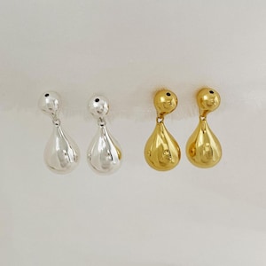 May include: Two pairs of teardrop earrings. One pair is silver, the other is gold. Each earring has a small ball at the top and a larger, rounded teardrop shape hanging below. The earrings are displayed against a white background.