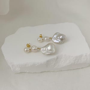 May include: A pair of pearl earrings. Each earring has three pearls, with the largest pearl in an irregular, organic shape. The earrings are set on gold posts and are displayed on a white, textured surface.