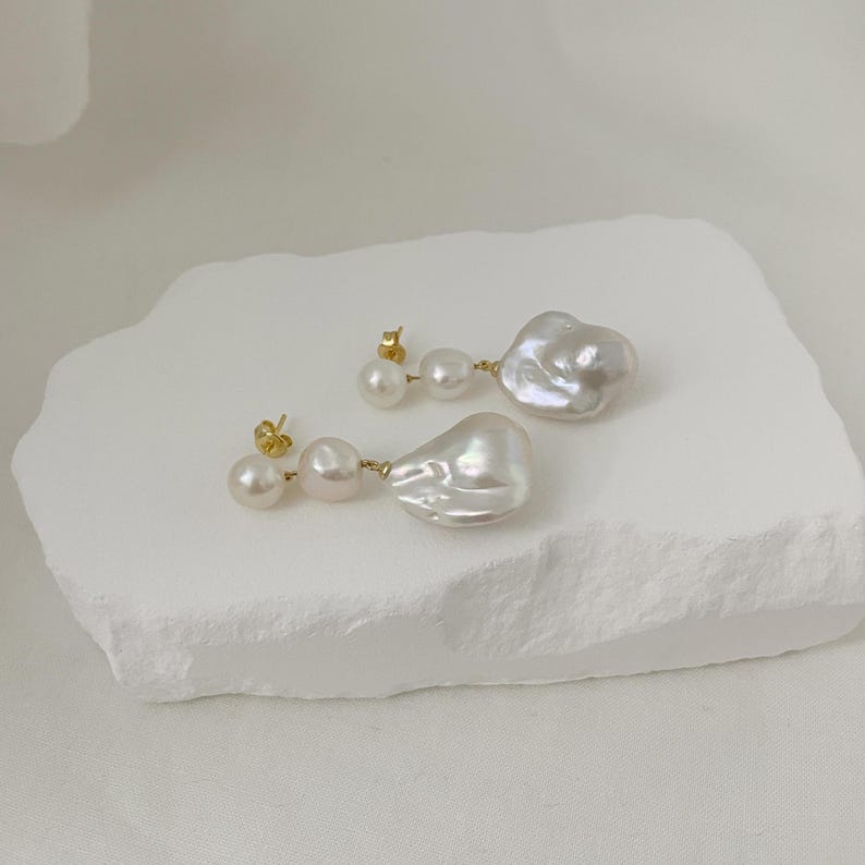 May include: A pair of pearl earrings. Each earring has three pearls, with the largest pearl in an irregular, organic shape. The earrings are set on gold posts and are displayed on a white, textured surface.
