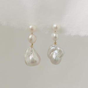 Op de afbeelding: Een paar elegante oorbellen met elk drie witte parels. De bovenste twee parels zijn rond, terwijl de onderste parel een grote, onregelmatige barokparel is. De oorbellen hebben goudkleurige accenten en hangen aan een delicate gouden draad.