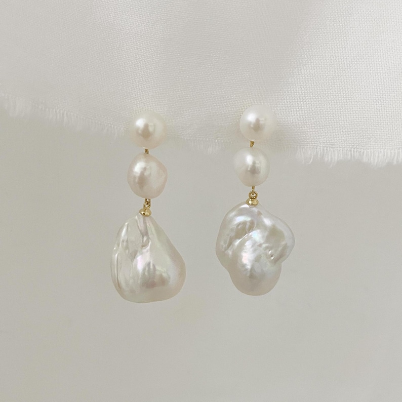 May include: A pair of earrings with three white pearls each. The top two pearls are round, while the bottom pearl is a large, irregular baroque pearl. The earrings have gold-coloured accents and hang from a delicate gold wire.