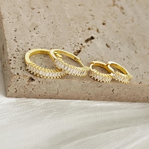 May include: Three pairs of gold hoop earrings with a row of clear baguette-shaped cubic zirconia stones. The earrings are marked "S925" on the back.