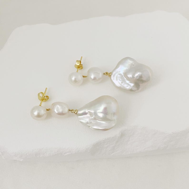 May include: A pair of earrings with three white pearls each. The earrings have a gold-coloured post and connecting wire. The largest pearl has an irregular, baroque shape.