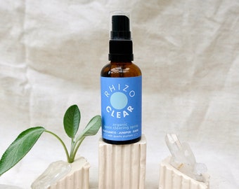 May include: A brown glass bottle with a blue label that reads "RHIZO CLEAR organic space clearing spray palo santo - juniper - sage with quartz crystals" and a black spray nozzle. The bottle is sitting on a white ceramic stand with a sage plant in a white ceramic pot next to it. There are several clear quartz crystals on the white surface.