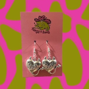 May include: Silver heart-shaped earrings with devil horns and a tail. The earrings hang from silver hooks and are displayed on a pink card with a frog wearing a cowboy hat graphic. The background is pink and green.