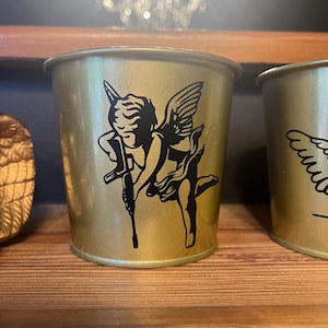 May include: A gold metal cup featuring a black silhouette of an angel holding a gun. The cup is on a wooden surface.
