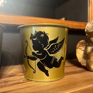 May include: A gold metal cup featuring a black silhouette of a cupid holding an arrow and wings. The cup is on a wooden surface.