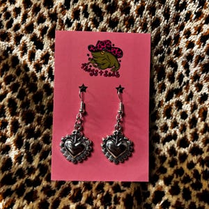 May include: Silver heart earrings with a decorative border and a cross design. The earrings hang from a small chain and a star-shaped hook. The earrings are displayed on a pink card with a frog logo that says "Froggs + Sods".