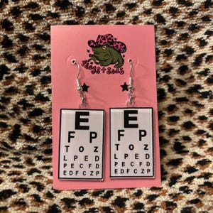 May include: Eye chart earrings with silver hooks. Each earring features a white background with black letters, spelling out an eye chart. The earrings are attached to a pink card with a frog wearing a cowboy hat.