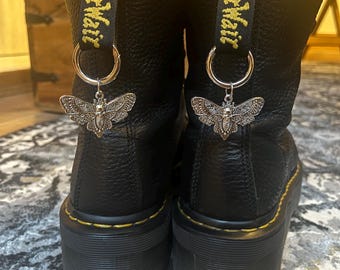 Puede incluir: Botas de plataforma de cuero negro con costuras amarillas y presillas "AirWair". Cada bota tiene un dije de plata con forma de polilla colgando de un anillo. Las botas tienen un aspecto texturizado y una suela gruesa.