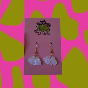 May include: A pair of white cloud-shaped earrings with gold hooks. The earrings are on a pink card with a cartoon frog wearing a cowboy hat and the text "Frogs + Sass".