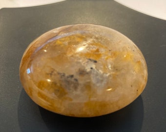 May include: A smooth, round, yellow-brown gemstone with white and black inclusions. The stone is polished and has a glossy finish.
