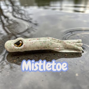 May include: A white and green fishing lure with red speckles and the word "Mistletoe" in a blue and white font. The lure has a gold-colored eye and multiple tentacles. The lure is lying in water.
