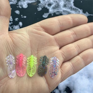 May include: Five colorful fishing lures are displayed in a hand. The lures are in shades of clear, pink, green, brown, and purple. Each lure has a segmented body and a tail with small, decorative elements. The background is a blurred view of a snowy surface.