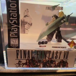 May include: A clear plastic cube display case with a miniature diorama of the video game Final Fantasy VII. The diorama features a character holding a large sword, and the game's logo is visible on the base of the display case.