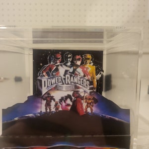 May include: A clear acrylic cube display case containing a Power Rangers The Movie poster. The poster shows the five Power Rangers in their signature suits, standing on a snowy mountain. The base of the case features a Power Rangers logo.