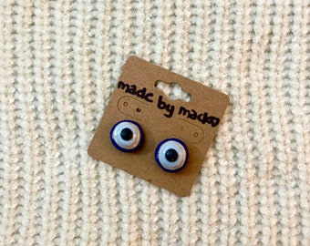 May include: A pair of blue and white evil eye earrings on a brown card with the text "made by made" on it.