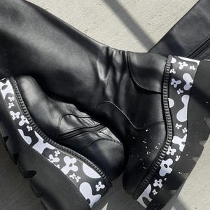 Könnte beinhalten: Schwarze, kniehohe Stiefel aus Leder mit einer dicken Plateau-Sohle. Die Plateau-Sohle hat ein weiß-schwarzes abstraktes Muster.