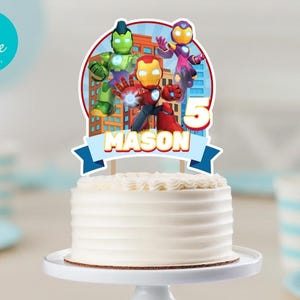Puede incluir: Una tarta blanca con un adorno tem&aacute;tico de superh&eacute;roes. El adorno presenta a Iron Man y otros personajes, el n&uacute;mero 5 y el nombre "Mason" en una pancarta azul. La tarta est&aacute; sobre un soporte blanco.