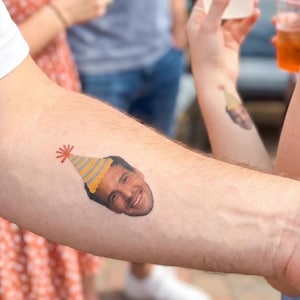 May include: A temporary tattoo applied to the skin featuring a realistic photographic portrait of a smiling person wearing a yellow and grey striped party hat with a red pom-pom on top. The design is printed in full color with clear details.