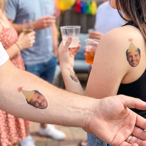 May include: Temporary tattoos of a smiling face wearing a party hat are visible on the arm and shoulder of several people. The tattoos are in color and appear to be applied to the skin.