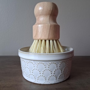 May include: A white ceramic dish brush with a wooden handle and natural bristles. The brush sits in a decorative, round, white ceramic holder with a dotted pattern. The wooden handle is light-colored and rounded.