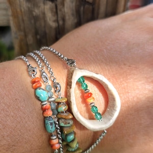 May include: A multi-strand bracelet featuring turquoise, coral, and stone beads, along with a pendant made from a bone with green, orange, and green beads. The bracelet is on a silver chain.