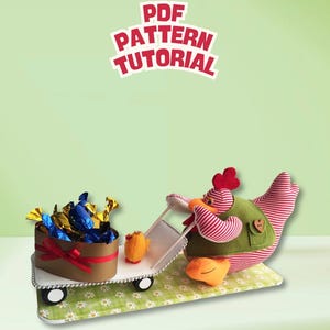 Puede incluir: Un pollo de peluche con rayas rojas y blancas y un chaleco verde tirando de un carrito blanco. El carrito contiene una caja marrón en forma de corazón llena de dulces envueltos en azul y oro. El texto "PDF PATTERN TUTORIAL" aparece en rojo.