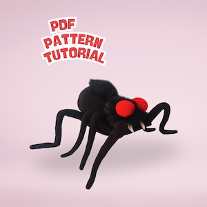 Puede incluir: Una araña negra de peluche con grandes ojos rojos y largas patas curvas. La araña tiene un cuerpo peludo y está sobre un fondo rosa. El texto en la esquina superior izquierda dice "PDF PATTERN TUTORIAL".