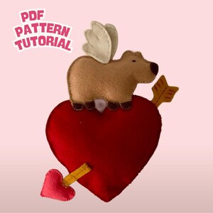 Puede incluir: Decoración de San Valentín hecha a mano de fieltro con un carpincho marrón con alas color crema sobre un corazón rojo atravesado por una flecha amarilla. Un corazón rojo más pequeño está unido a la flecha. El texto "PDF PATTERN TUTORIAL" está arriba a la izquierda.