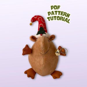 Puede incluir: Una figura de peluche marrón con forma de gnomo con un gorro rojo y un pequeño árbol de Navidad en la cabeza. La figura tiene pequeños brazos y piernas marrones y sostiene una campana dorada. El texto superior dice "PDF PATTERN TUTORIAL".