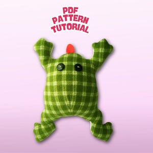 Peut inclure: Un jouet en peluche vert à carreaux avec des yeux en bouton et une corne rouge. Le jouet est dans une pose de grenouille avec les bras et les jambes tendus. Le texte "PDF PATTERN TUTORIAL" est au-dessus du jouet.