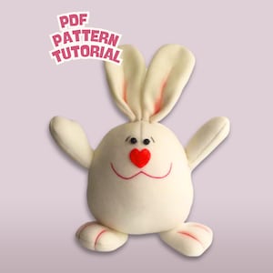 Puede incluir: Un peluche blanco con forma de conejito con una nariz roja en forma de corazón y una boca sonriente. El conejito tiene orejas largas con el interior rosa, brazos extendidos y detalles rojos en los pies. El texto "PDF PATTERN TUTORIAL" es visible.
