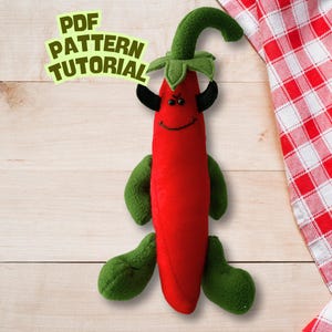Puede incluir: Un peluche de pimiento rojo con brazos, piernas y tallo verdes, y cuernos negros. El pimiento tiene una cara sonriente y el texto "PDF PATTERN TUTORIAL" en verde y amarillo. Un paño a cuadros rojos y blancos está en el fondo.