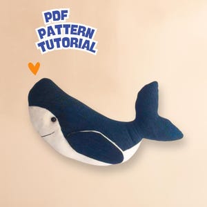 May include: A plush whale toy in navy blue and white, with a smiling face and a curved tail fin. The image includes the text "PDF PATTERN TUTORIAL" and a small orange heart.