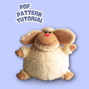 Puede incluir: Un peluche de oveja con cuerpo color crema, orejas marrones y nariz en forma de corazón rojo. La oveja tiene una cara sonriente y está sobre un fondo azul claro. El texto "PDF PATTERN TUTORIAL" aparece en la parte superior.