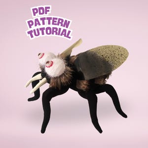 Op de afbeelding: Een pluche speelgoedvlieg met grote, wiebelende ogen en een harig lichaam. De vlieg heeft zwarte poten en vleugels, en de tekst "PDF PATTERN TUTORIAL" staat erboven. De vleugels zijn een mix van zwart en beige.
