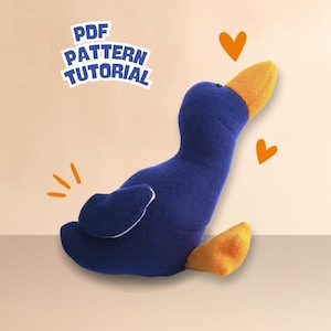 Könnte beinhalten: Eine blaue Plüschente mit orangefarbenem Schnabel und Füßen, mit dem Text "PDF PATTERN TUTORIAL". Die Ente sitzt, mit orangefarbenen Herz- und Burst-Grafiken.