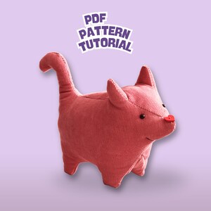 Puede incluir: Un gato de peluche de pana de color rosa rojizo, con una nariz roja en forma de corazón y ojos de botón negros. El gato tiene una cola larga y orejas puntiagudas. La imagen incluye una pegatina que dice "PDF PATTERN TUTORIAL".